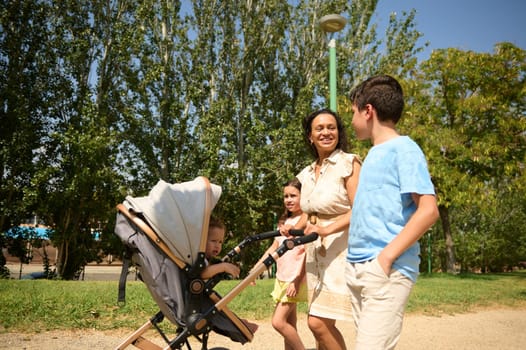 A joyful family strolls through a sunlit park, pushing a stroller. They enjoy quality time together surrounded by nature and greenery, smiling under the clear blue sky.