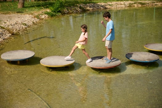 Two children balance on round stepping stones over a clear, shallow pond in a park setting.