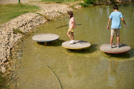 Two children playfully balance on circular platforms above shallow water in a sunny park setting, showcasing fun and adventure in an outdoor environment.