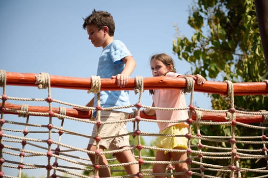 Two young children playing on a rope bridge in an outdoor park. The scene captures joy, exploration, and the spirit of adventure against a backdrop of trees and a clear blue sky.