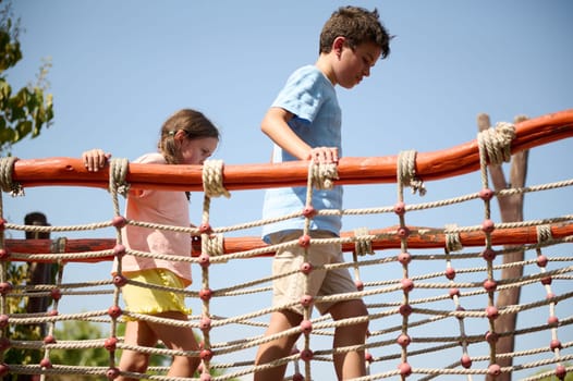 Two children carefully walk across a rope bridge in an outdoor playground. The scene is bright and emphasizes a sense of adventure and playfulness under a clear sky.
