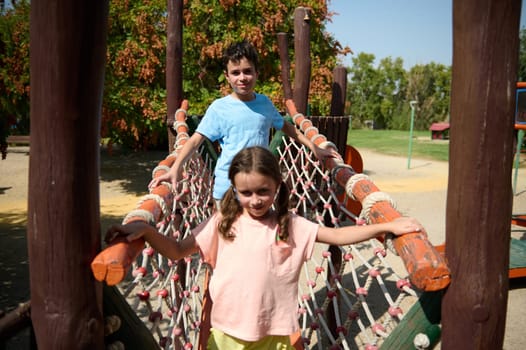 Two children enjoy a sunny day playing on a rope bridge at the park. Laughter and adventure fill the air as they explore the playground together.