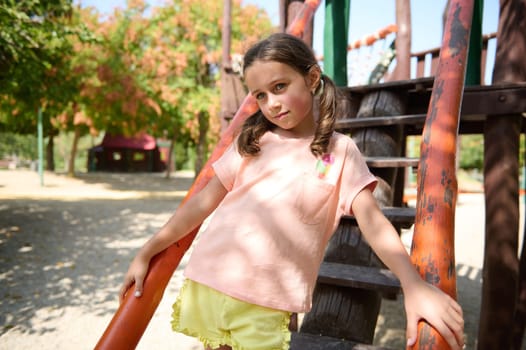 A child in casual clothing poses confidently on playground equipment surrounded by trees.