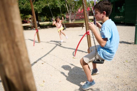 Two children enjoying a sunny day on swings at a park. The boy and girl are smiling and having fun, capturing the essence of carefree childhood joy.