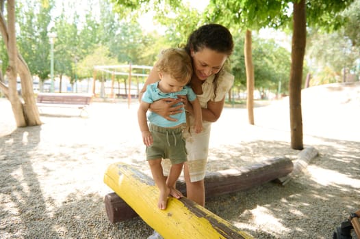 A mother supports her young child on a balance beam in a sunlit park, fostering a sense of adventure and trust in an outdoor family environment.
