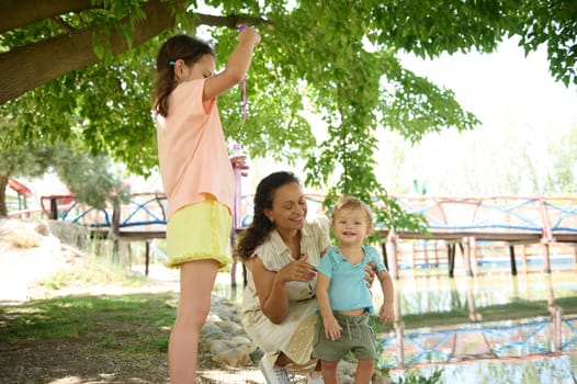 A joyful mother plays with her two children beneath lush greenery by a pond, fostering family bonds and enjoying nature.