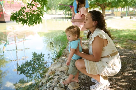 A mother and her child explore nature near a serene pond, surrounded by greenery. The peaceful scene captures a moment of bonding and curiosity in a natural setting.