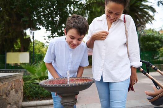 A joyful mother and son smile together as they interact with a park fountain on a sunny day.