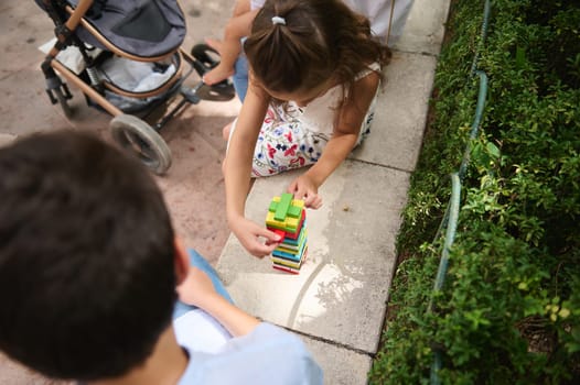 A young girl is engaged in building with colorful blocks outdoors next to a stroller. The setting features lush greenery, creating a playful and creative atmosphere.