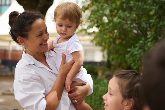 A loving mother holds her baby while enjoying a sunny day with family outdoors. Smiles and affection create a warm and joyful scene, symbolizing family love and connection in a natural setting.