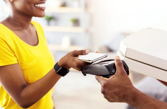 Unrecognizable Black Lady Paying Courier Using Mobile Phone As Credit Card Putting Cellphone To Payment Terminal Receiving Pizza Boxes At Home. Cropped, Panorama