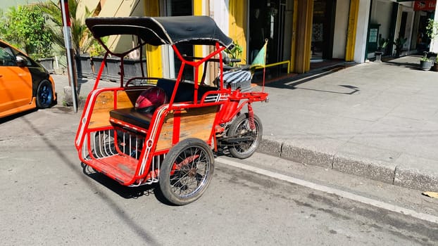 pedicab or rickshaw or locals calls it becak with a canopy is parked on a sunny street, reflecting a vibrant urban scene. The vehicle features a padded seat and unique design