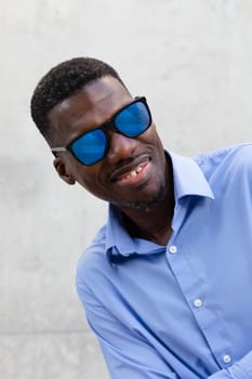 Smiling African American Man In His 40s With Blue Sunglasses And Shirt. Portrait Of Happy Stylish Middle Age Male, Gray Concrete On Background. Accessories. Vertical Plane. High quality photo