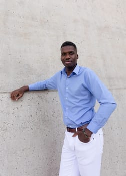 Portrait Of Handsome Smiling Tall African American Man wearing White Jeans and Blue Shirt, Brown Belt Outdoor, Concrete Wall on Background. Copy Space Fashionable Male, Ivorian. Vertical.