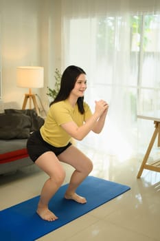 Smiling young woman doing squats during home workout. Fitness and healthy living concept.
