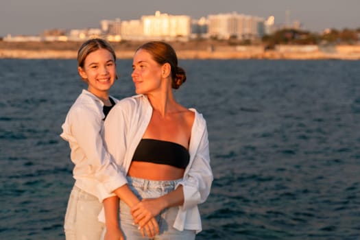 Two young women, one in a white shirt and the other in a black bikini top and jeans, stand by the ocean at sunset, holding hands and looking at each other.