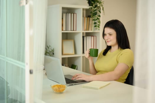 Happy young woman working on her laptop while enjoying a cup of coffee in organized home.