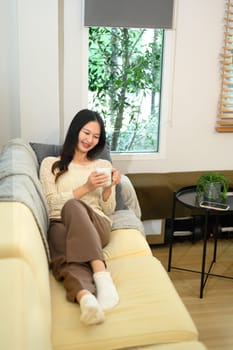 Happy asian woman relaxing on couch at home and sipping a hot beverage.