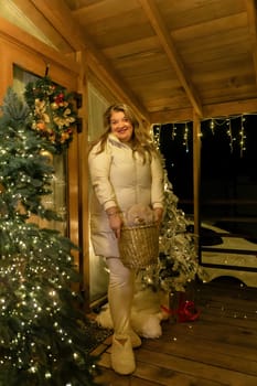 A woman in a white fuzzy coat and slippers stands beside a decorated Christmas tree in a wooden cabin. She smiles and holds a basket.