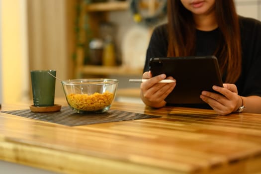 Young woman using digital tablet at kitchen table with bowl of cereal and a glass of water.