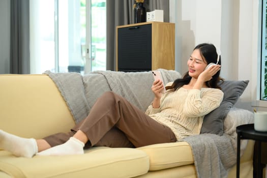 Relaxed young asian lady in wireless headphones using smartphone on sofa.