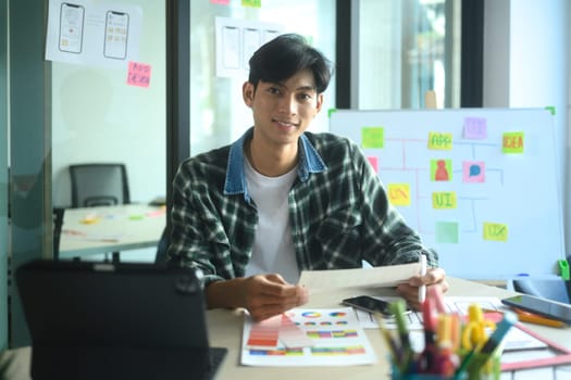 Confident young designer working in modern office surrounded by tools and sketches for UI UX design.