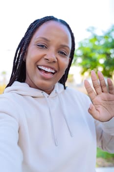 Young Woman with braided hair wearing a hoodie, smiling brightly while waving at the camera. She appears to be outdoors on a sunny day, enjoying a video call or taking a selfie with greenery in the background.