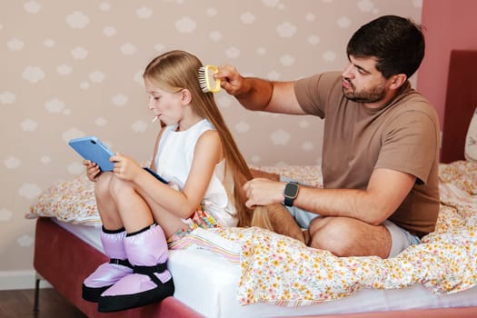 A father and daughter sit on a bed. The father is brushing the daughter's long hair. She is looking at a tablet and holding a lollipop. The bed has a floral patterned duvet.