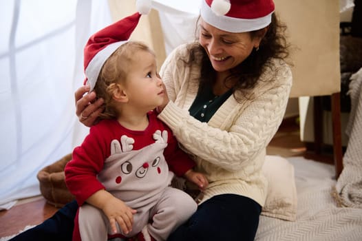 A joyful mother and child share a festive moment indoors, both wearing Santa hats, creating warm holiday memories together.