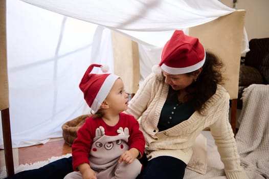 A joyful mother and her child wearing Santa hats share a playful holiday moment indoors.