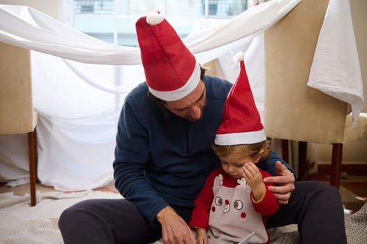 A father and child share a tender moment in an indoor fort, wearing Santa hats. The scene captures holiday joy and the warmth of family bonding during the festive season.