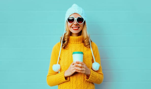 Winter portrait smiling young woman with coffee cup wearing yellow knitted sweater, white hat, glasses on blue background