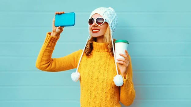 Winter portrait smiling woman taking selfie picture by phone with coffee cup wearing yellow knitted sweater, white hat, heart shaped sunglasses on blue background