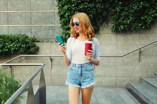 Stylish smiling young woman using smartphone with coffee cup in the city