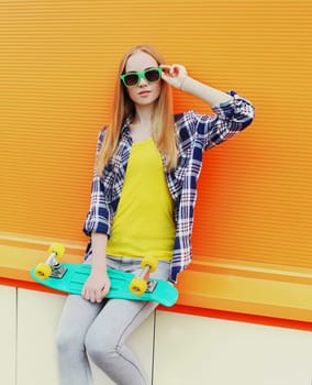 Fashion pretty blonde girl wearing a sunglasses with skateboard over colorful orange background