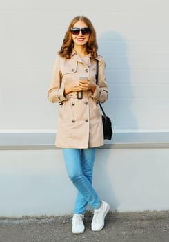 Fashion lovely young smiling woman using smartphone wearing a coat handbag clutch in city