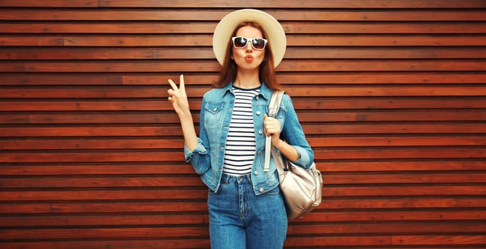 young woman blowing kiss with phone in summer hat, backpack and jean jacket on wooden background