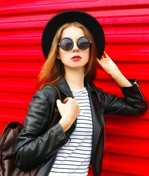 Fashion portrait young woman in black rock style hat, jacket stands with backpack on red background