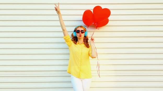 Happy young woman with bunch of red heart shaped balloons listening to music in headphones and blowing kiss on white background