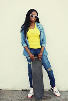 Full length stylish smiling young african woman posing with skateboard in jeans denim clothes in the city