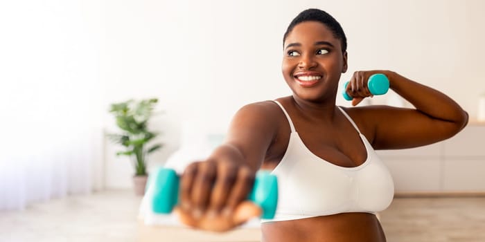 Home sports for weight loss. Happy plus size black woman doing fitness exercises with dumbbells indoors. Millennial overweight lady strengthening her biceps, leading active lifestyle