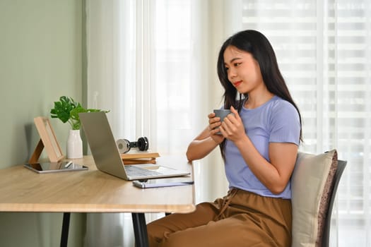 Pretty young woman drinking herbal tea and reading email on laptop screen.