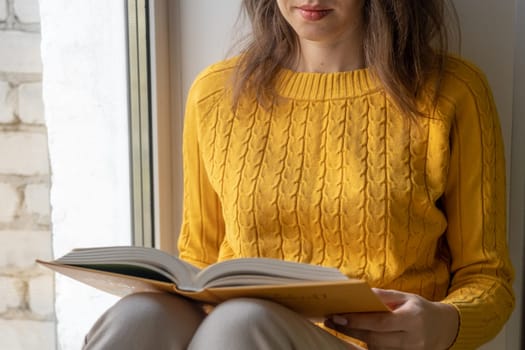 Young beautiful woman sitting by the window yellow knitted sweater read book, daily planner, notepad. Relax concept. Hold cappuccino glass of coffee with white foam. Text is out of focus