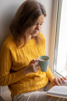 Young beautiful woman sitting by the window yellow knitted sweater read book, daily planner, notepad. Relax concept. Hold cappuccino glass of coffee with white foam. Text is out of focus