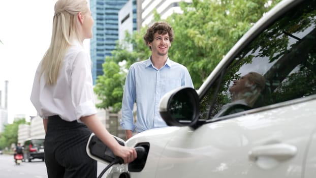 Progressive businessman and businesswoman install charger plug from charging station to electric car before driving around city center. Eco friendly rechargeable car powered by clean energy.