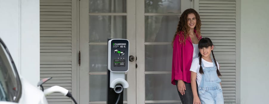 Happy little young girl learn about eco-friendly and energy sustainability as she help her mother recharge electric vehicle from home EV charging station. EV car and modern family. Panorama Synchronos