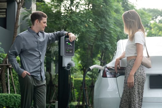 Young couple travel with EV electric car charging in green sustainable city outdoor garden in summer shows urban sustainability lifestyle by green clean rechargeable energy of electric vehicle innards