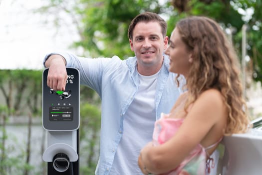 Happy and lovely couple with eco-friendly conscious recharging electric vehicle from EV charging station. EV car technology utilized as alternative transportation for future sustainability. Synchronos