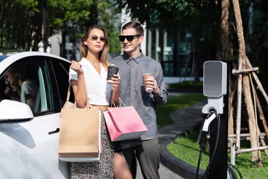 Young couple travel with EV electric car charging in green sustainable city outdoor garden in summer shows urban sustainability lifestyle by green clean rechargeable energy of electric vehicle innards