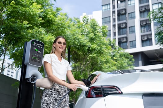 Young woman travel with EV electric car charging in green sustainable city outdoor garden in summer. Urban sustainability lifestyle by green clean rechargeable energy of electric BEV vehicle innards